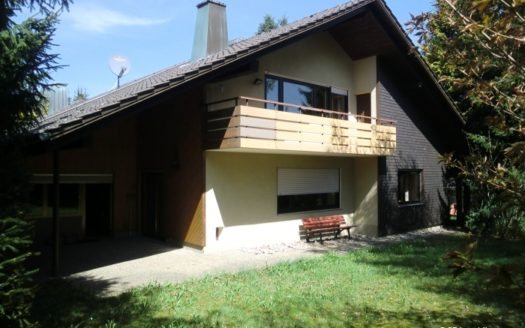 Hausansicht mit asymmetrischem Dach, Balkon und roter Bank